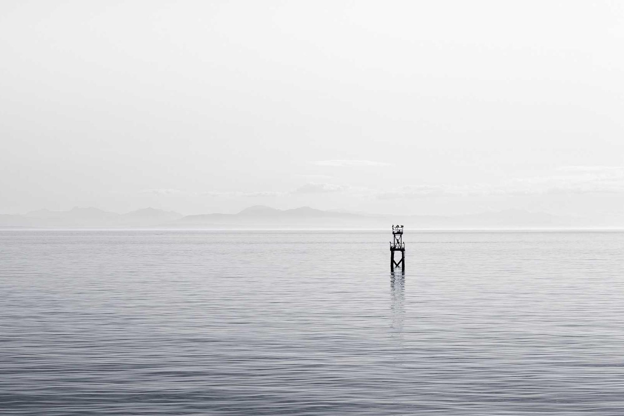 A still sea under a pale sky with one marker post in the water