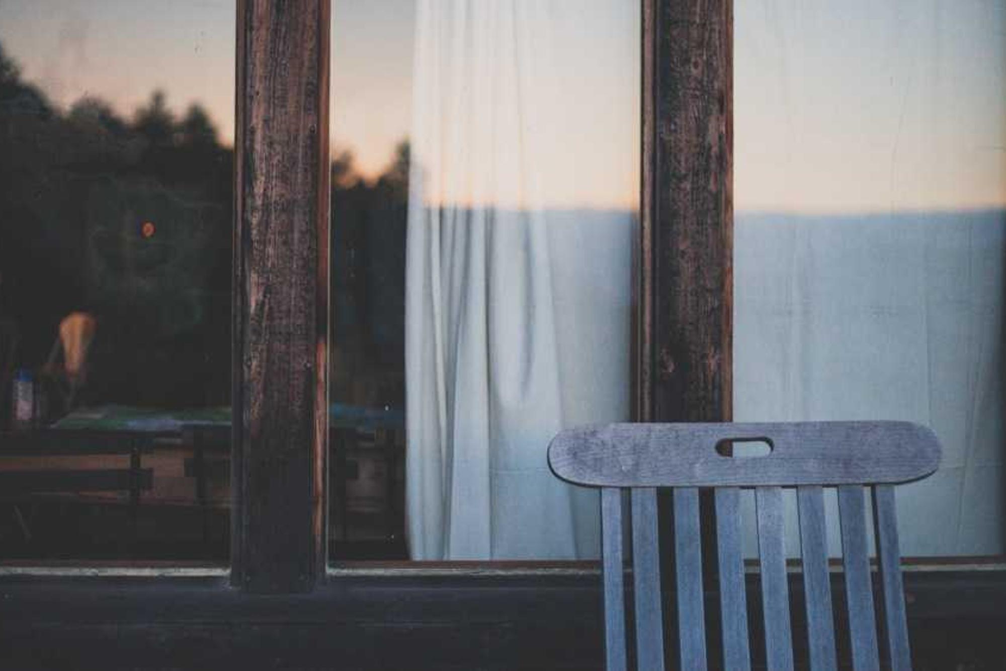 A wooden chair beside a window with a pale curtain at dusk